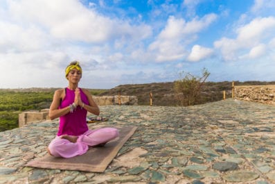 A lady in pink sitting in a yoga pose with her eyes closed on a clear and sunny day.